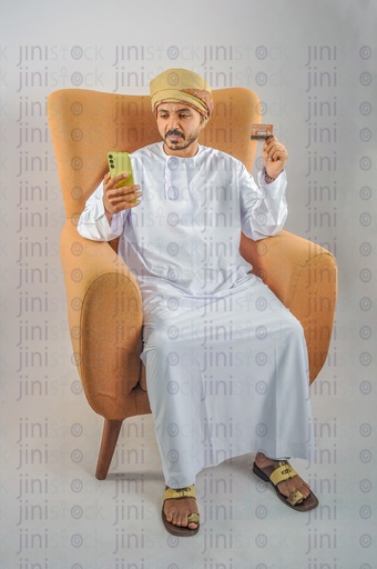 An Omani man wearing Omani uniform and sitting on a chair holding a mobile phone and visa card or card in his hands.