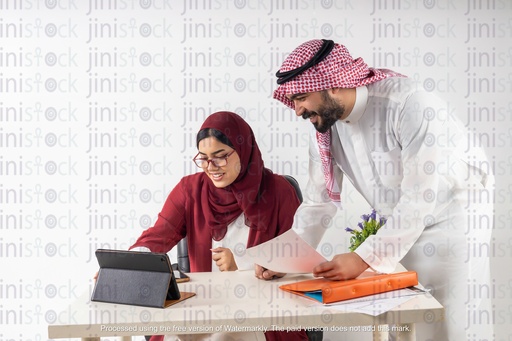 امرأة سعودية خليجية تجلس خلف مكتب ورجل خليجي يميل للتحقق من شيء ما على جهاز الآيباد